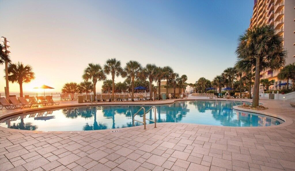 A swimming pool with lounge chairs and palm trees, surrounded by a hotel building.
