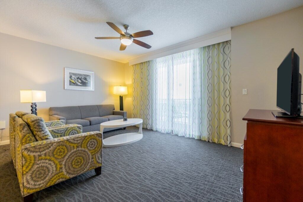A hotel room with a sofa, armchair, coffee table, TV, ceiling fan, and large windows with curtains.