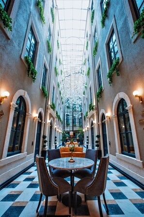 Un pasillo de hotel con piso a cuadros, ventanas arqueadas y una zona de estar central con una mesa redonda y sillas.