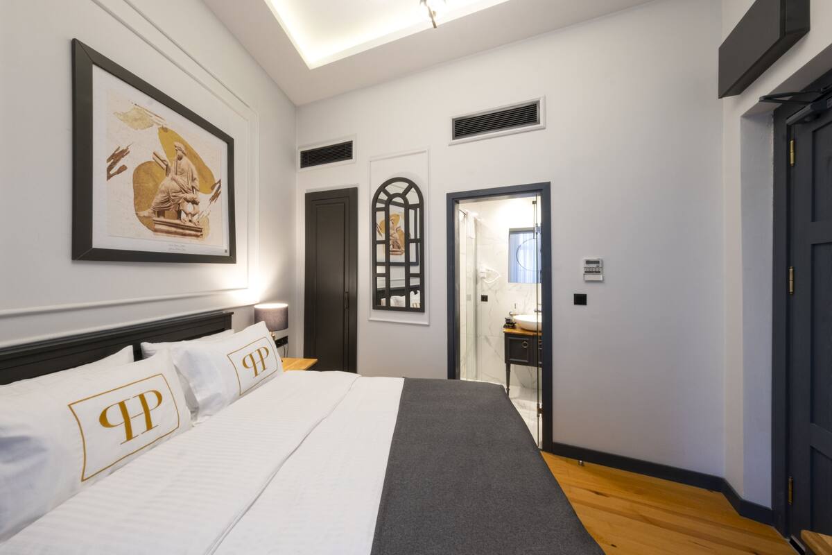 A modern bedroom with a large bed, a framed artwork above the bed, a dark door, and a bathroom visible through a glass door.