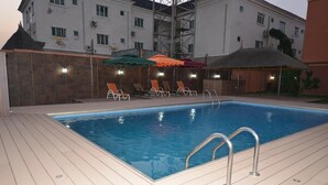 A swimming pool with lounge chairs and umbrellas, surrounded by a building with multiple windows and air conditioning units. - Elim Top Suites (Jos)