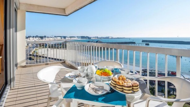 Un balcón con una mesa lista para el desayuno, con vista a una zona costera y una vista despejada del mar y edificios a lo lejos.