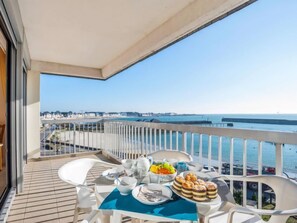 A balcony with a table set for breakfast, overlooking a coastal area with a clear view of the sea and distant buildings.
