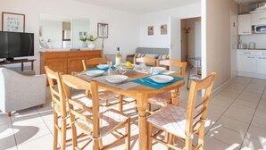 Una sala de estar moderna con una mesa de comedor preparada para cuatro personas, un sofá, un televisor y una zona de cocina con armarios y un microondas.