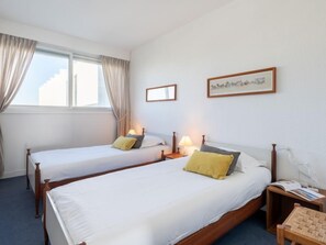 Une chambre d’hôtel avec deux lits simples, une petite table, une chaise, une table de chevet avec une lampe et une fenêtre avec des rideaux.