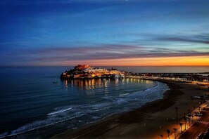 A coastal cityscape at dusk with a prominent fortified structure on a peninsula, a beach, and a promenade with palm trees.