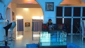 Amplia sala de estar con una mesa de centro de vidrio, un sofá y una mesa de comedor. Hay dos ventanas grandes con cortinas, y se ve una estatua a la izquierda.