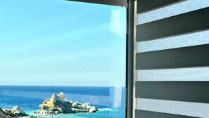A coastal view through a window with a patterned blind, a rocky shoreline, and a clear sky. - POPIS SEA VIEW APARTMENTS ¨MARIRENA 2¨ (Mades Maleviziou, Heraklion, Crete)