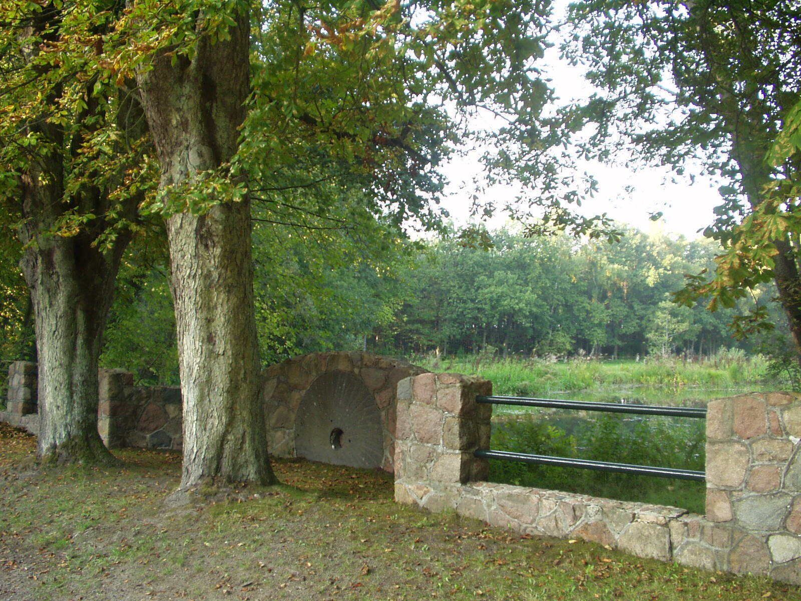 A stone wall with a metal railing, surrounded by trees and a grassy area.