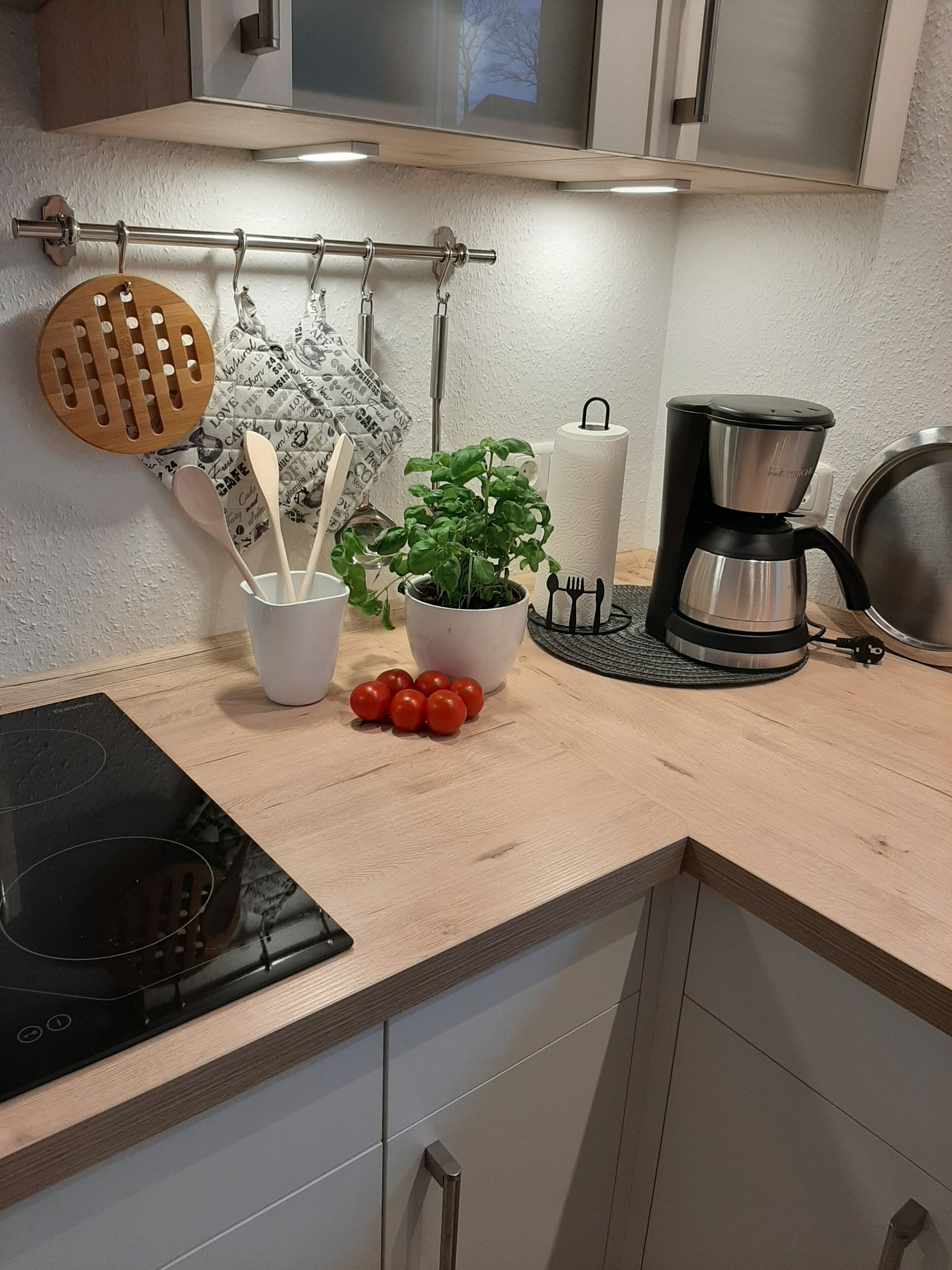 Eine Küche mit einer schwarzen Induktionskochplatte, einem hängenden Holz-Küchenhelferhalter, einem Topf mit Basilikum, einer Kaffeemaschine und einer Küchenpapierrolle.