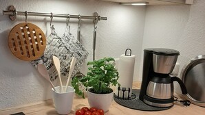 Una cucina con un piano cottura a induzione nero, un porta utensili in legno a sospensione, una pianta di basilico in vaso, una macchina per il caffè e un rotolo di carta assorbente.