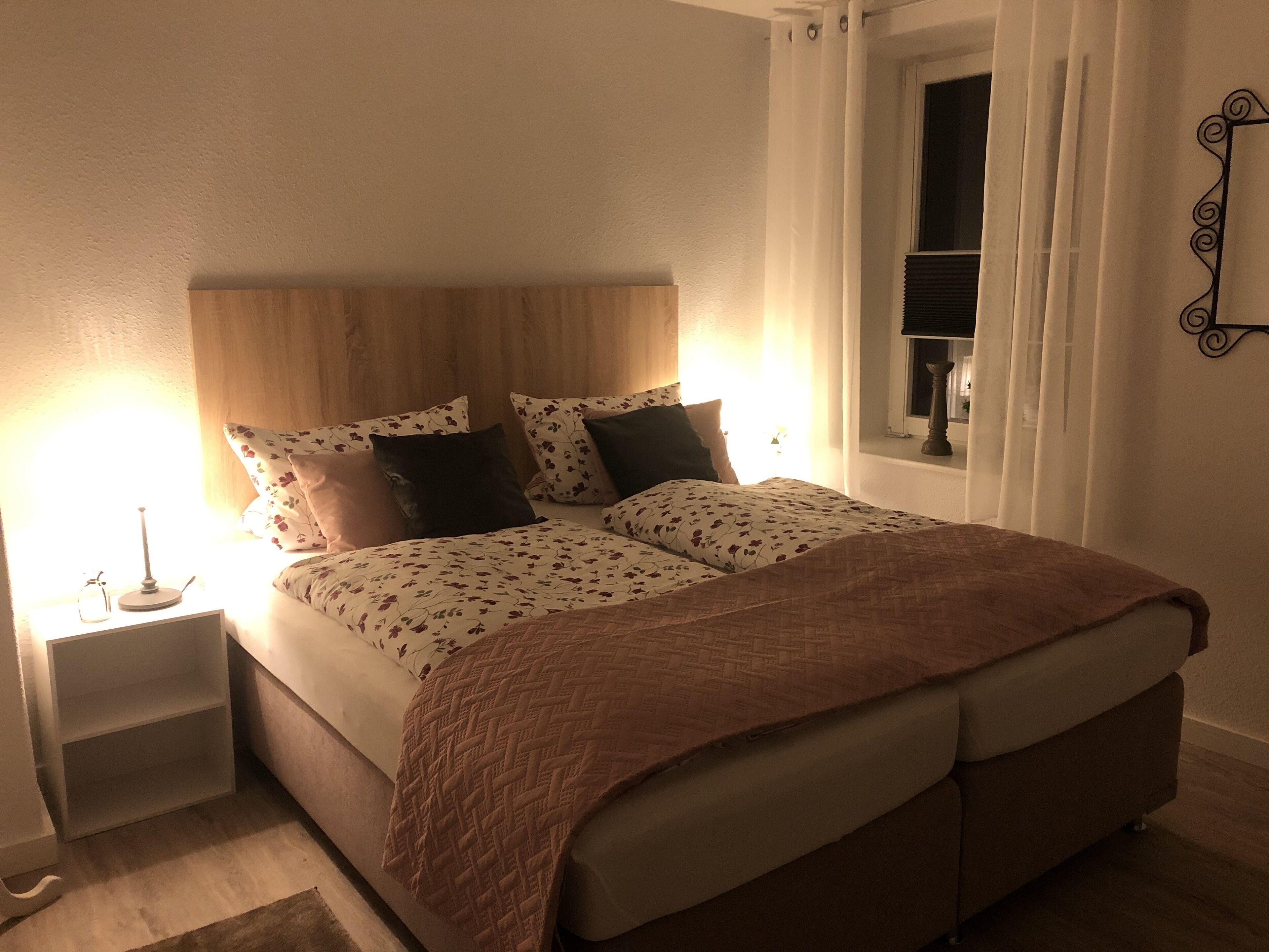 Ein ordentlich bezogenes Bett mit gemustertem Überwurf und Kissen, ein Nachttisch mit Lampe sowie ein Fenster mit Vorhängen.