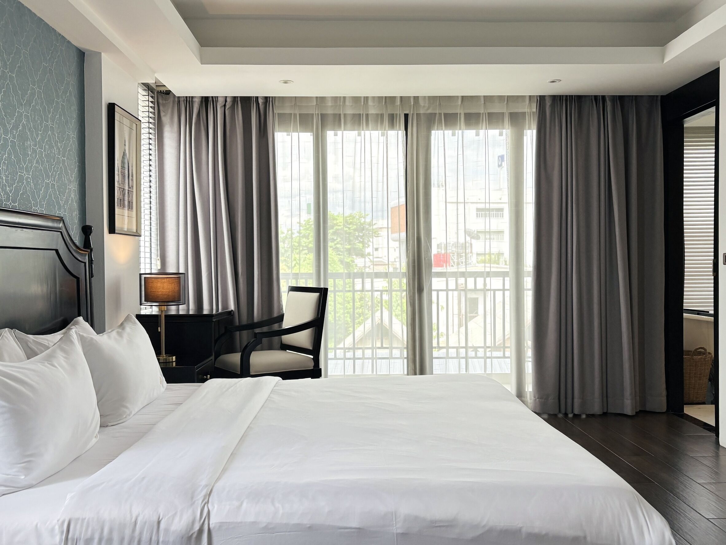 A hotel room with a large bed, a desk with a lamp, a chair, and a view of the outside through sheer curtains.
