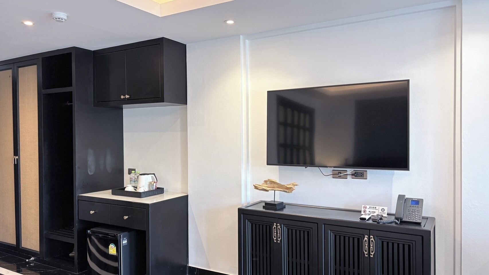 A modern hotel room with a flat-screen TV mounted on the wall, a small desk with a telephone, and a cabinet with a mirror.
