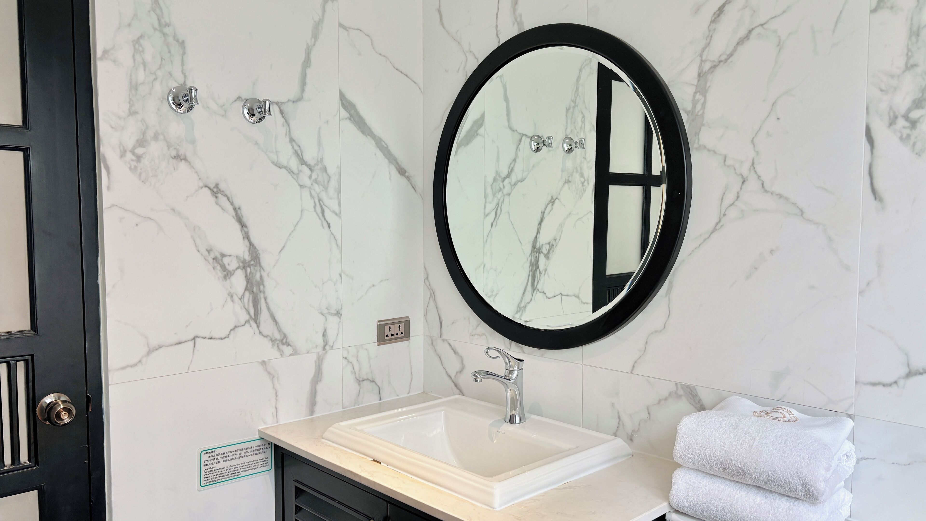 A bathroom with a marble wall, a round mirror, a sink, and a towel rack.