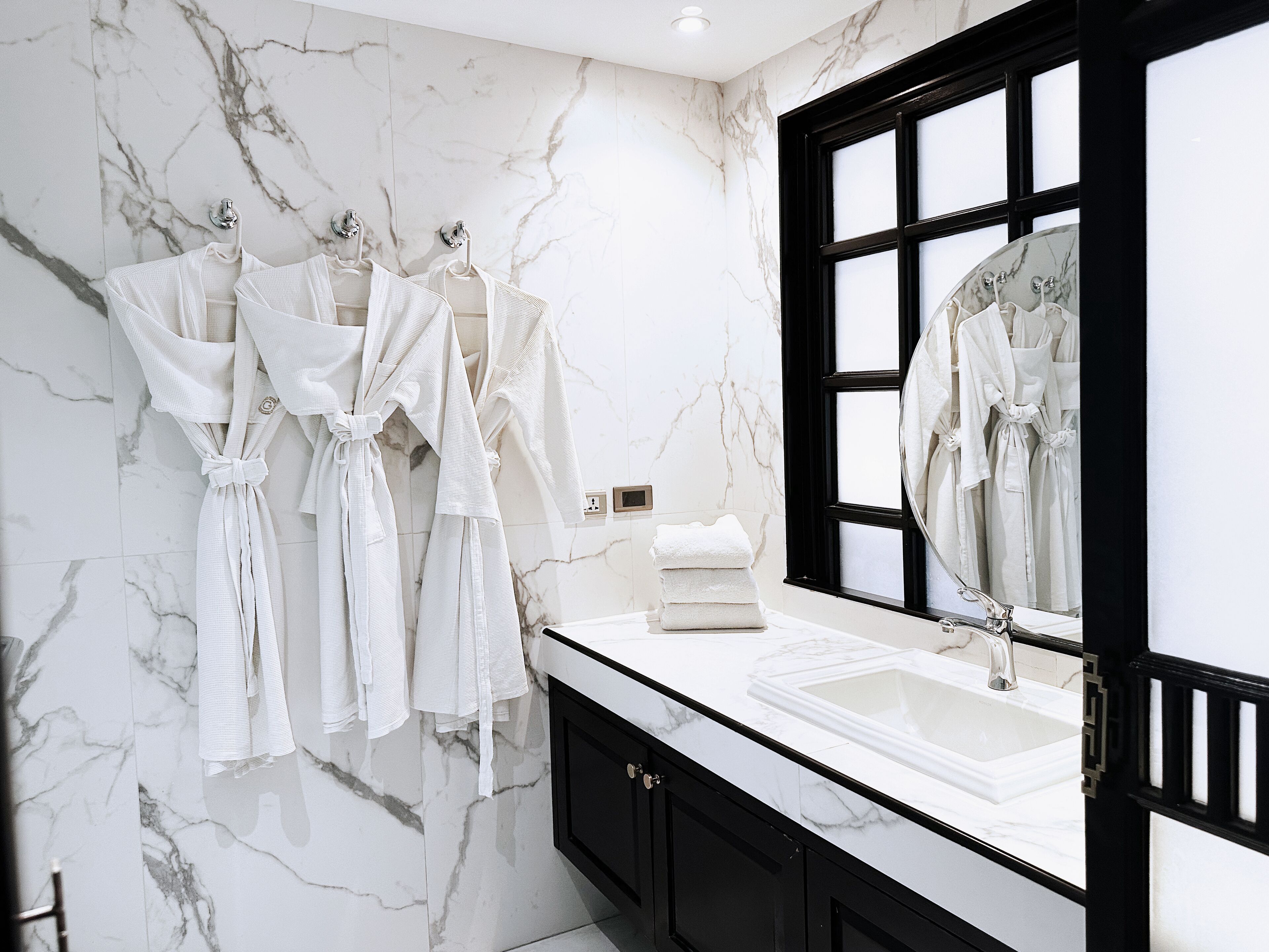 A bathroom with marble walls, a black-framed window, a white sink, and multiple white robes hanging on hooks.