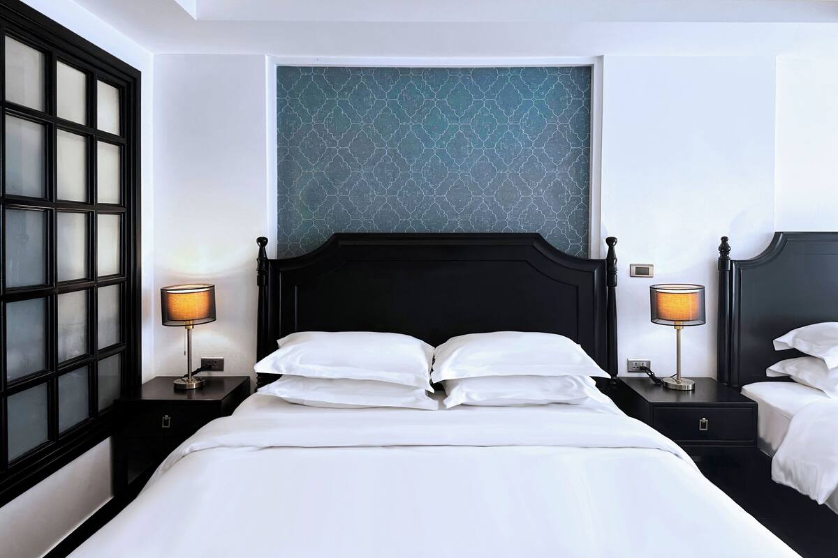 Habitación de hotel con una cama grande, dos mesitas de noche y una ventana con un diseño geométrico en la pared.