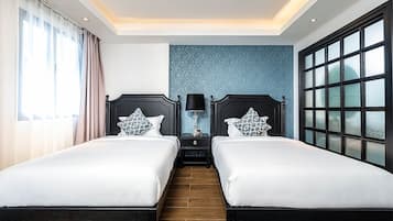 A hotel room with two beds, a nightstand with a lamp, a large window with blinds, and a blue accent wall.