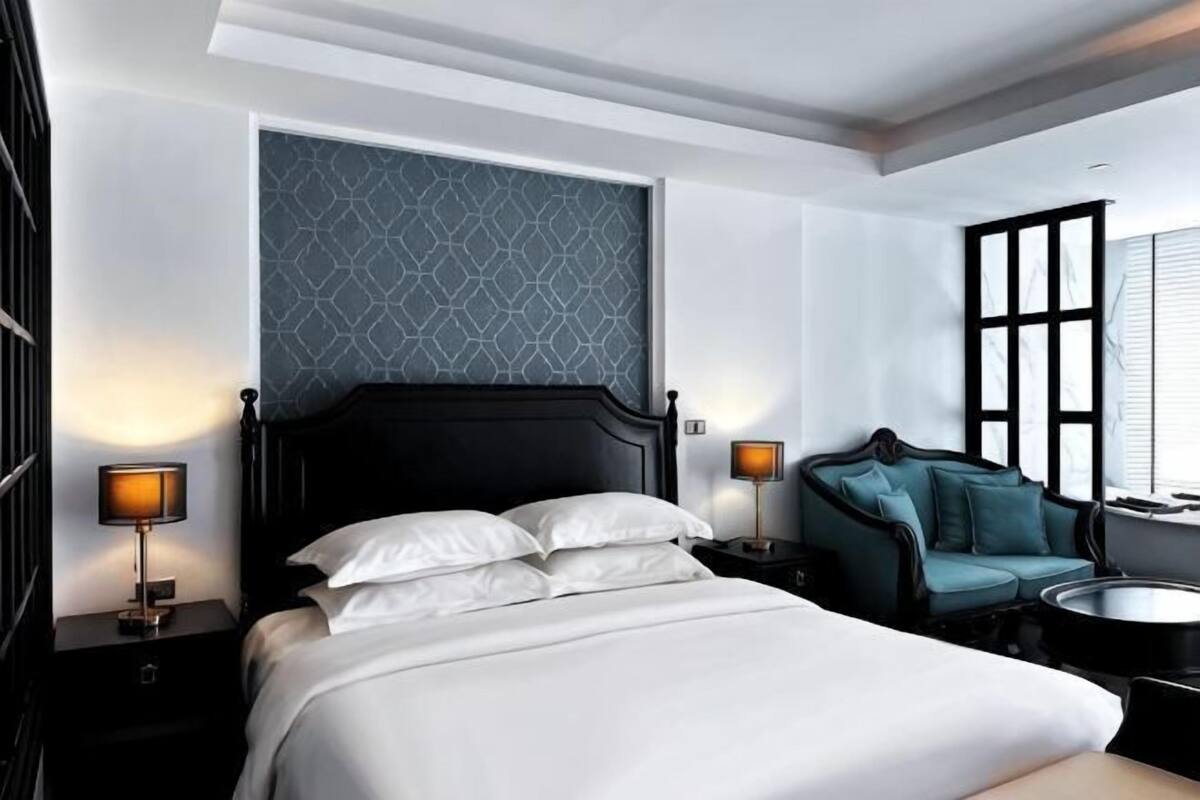 Una habitación de hotel con una cama grande, dos mesitas de noche con lámparas, un sofá y una ventana con persianas.