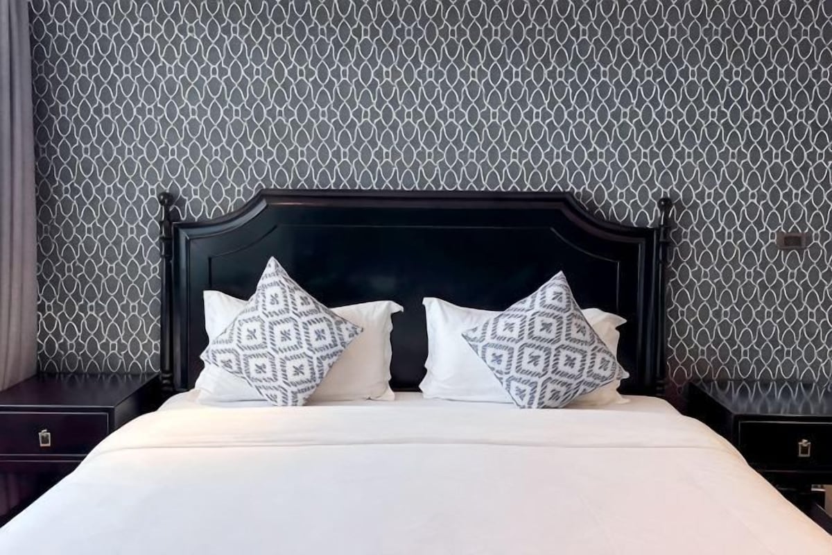 Una cama con cabecera oscura y almohadas estampadas, frente a una pared con diseño geométrico.