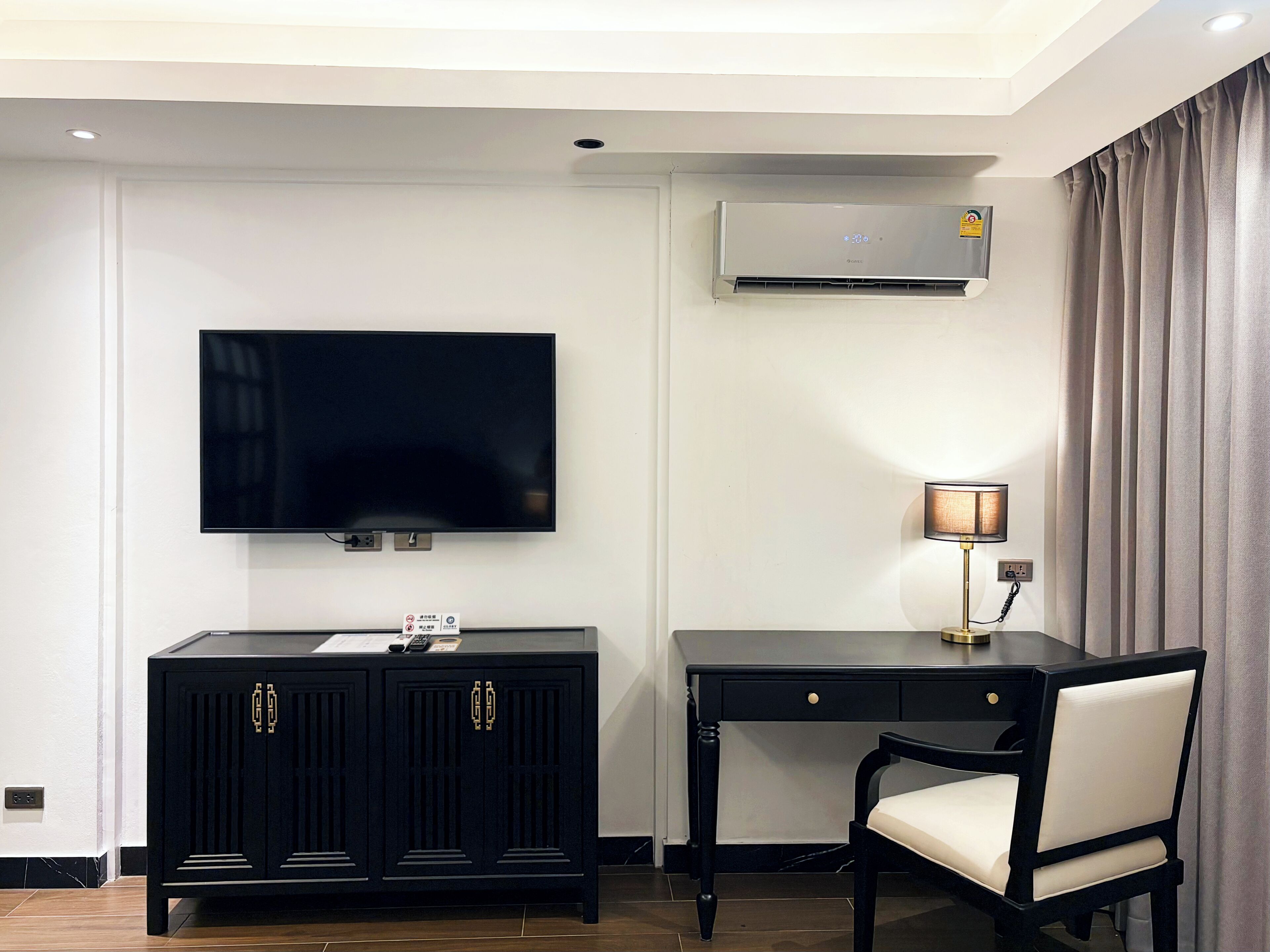 A hotel room with a flat-screen TV mounted on the wall, a desk with a lamp, a chair, and a dark-colored cabinet with drawers.