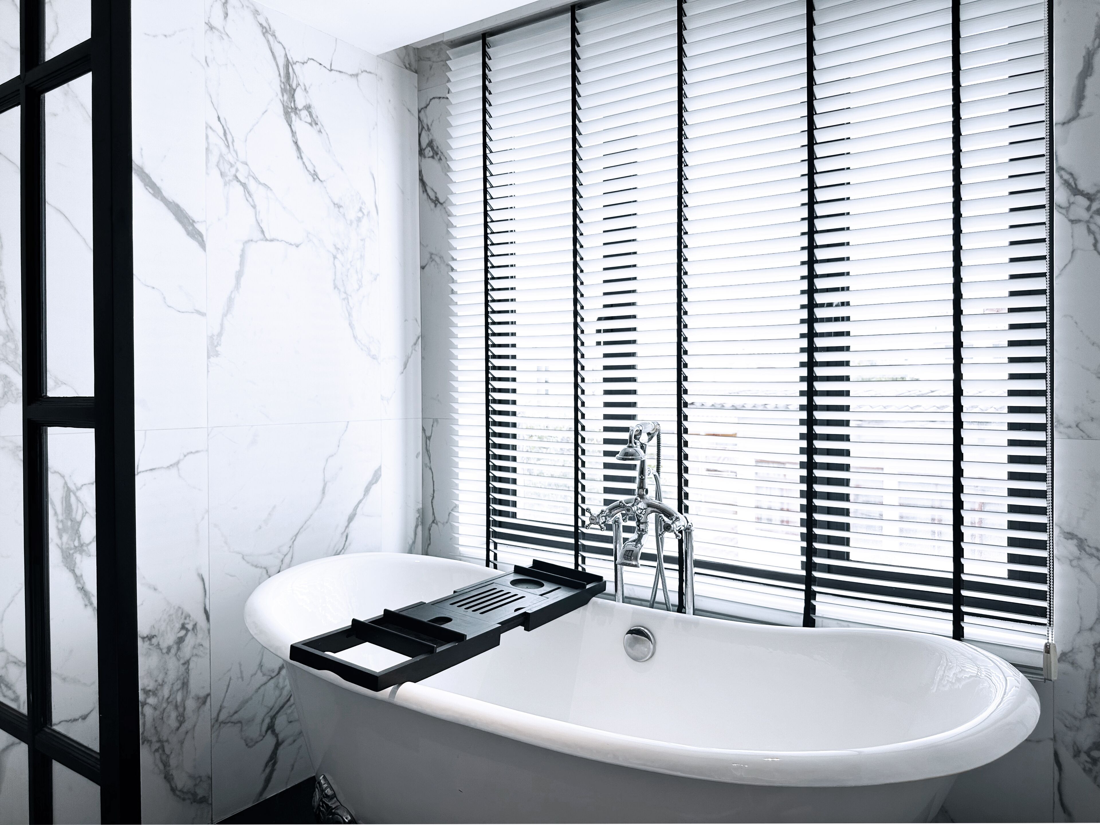 A modern bathroom with a freestanding bathtub, marble walls, and a window with blinds.