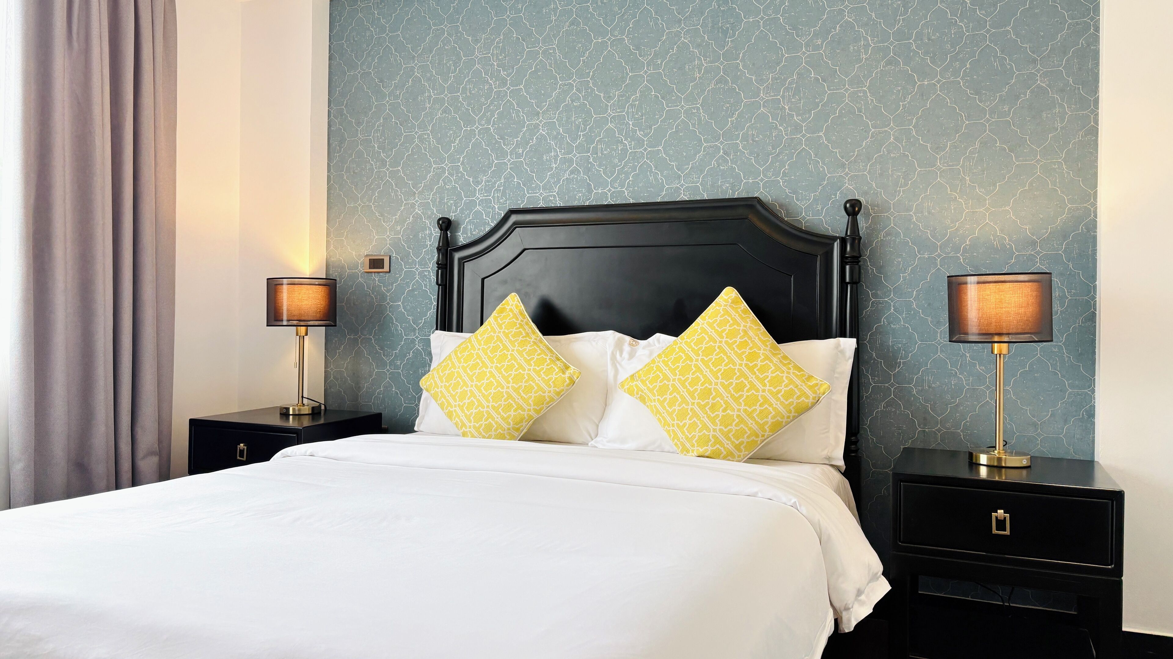A neatly made bed with white linens and two decorative pillows, flanked by nightstands with lamps, against a textured wall.