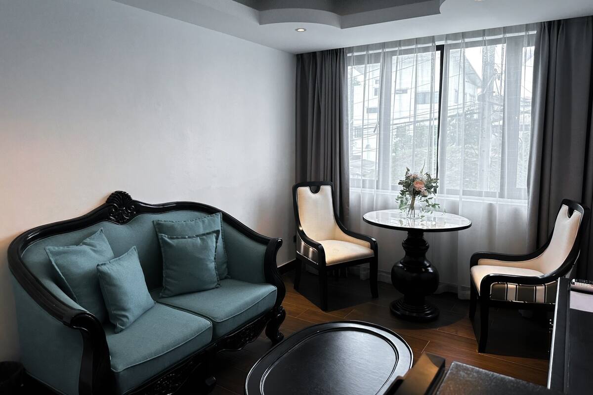 Una habitación con un sofá verde oscuro, dos sillas, una mesa redonda y un ventanal con cortinas.