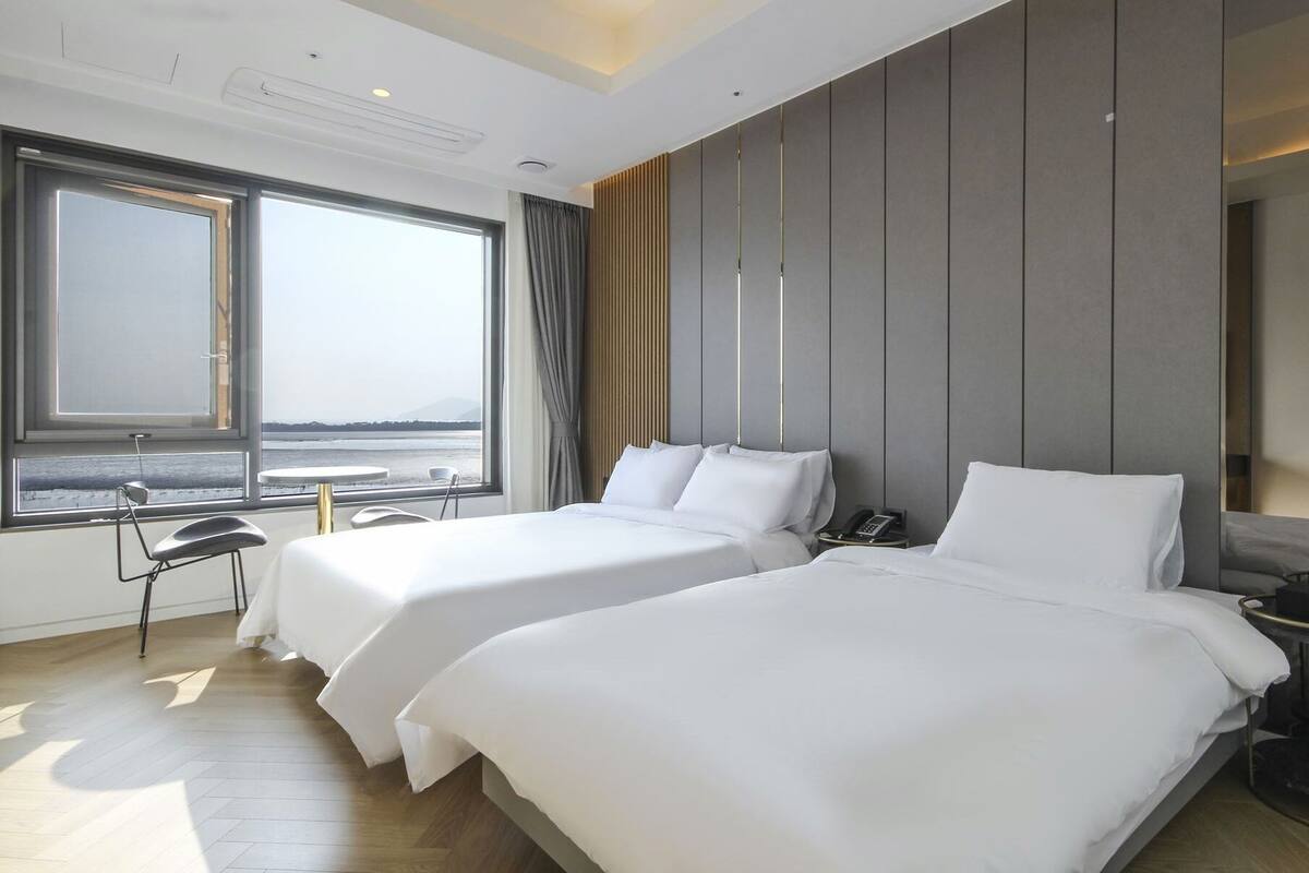 A hotel room with two beds, a desk, and a large window offering a view of the sea.