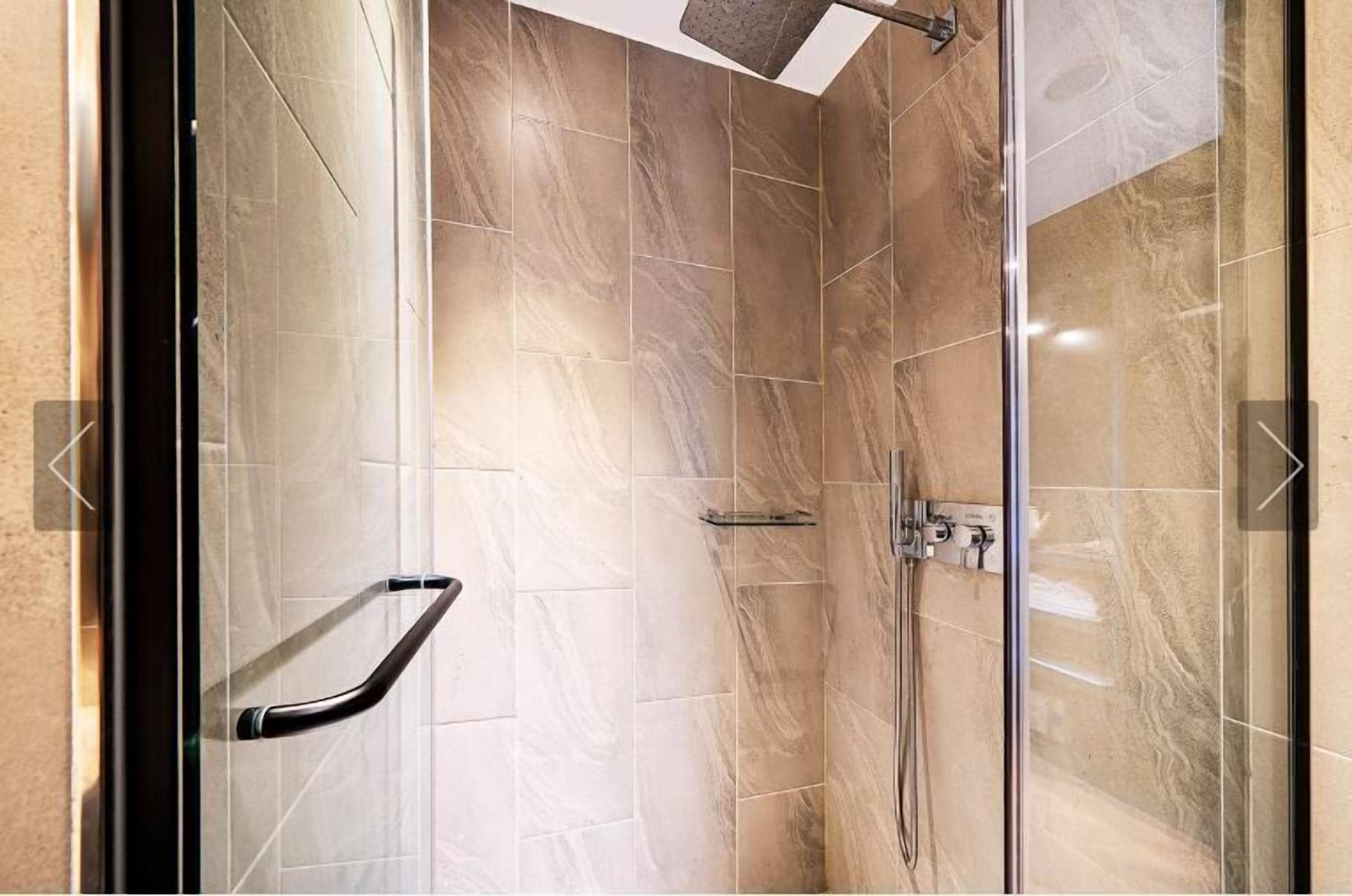 A glass-enclosed shower area with a handrail, a wall-mounted showerhead, and a control panel.