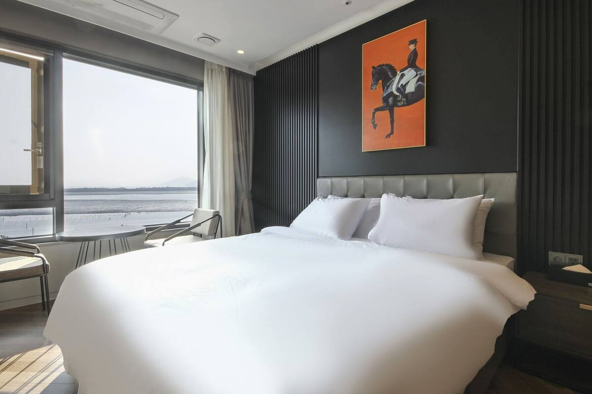 A hotel room with a large bed, a view of the sea, a modern chair, and a painting on the wall.