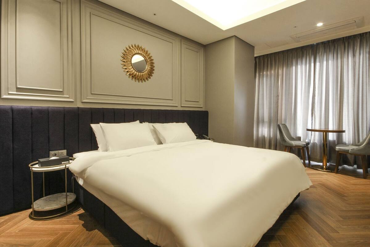 A hotel room with a large bed, a round mirror, two chairs, a small table, and a window with curtains.