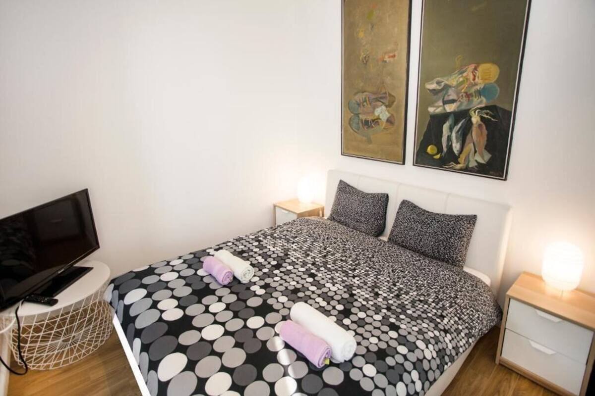 A neatly made bed with a polka dot bedspread, two pillows, and rolled towels. There is a television on a stand to the left and a bedside table with a lamp to the right.