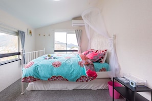 A bedroom with a bed, a window, an air conditioner, a bedside table, and a pink trash bin.