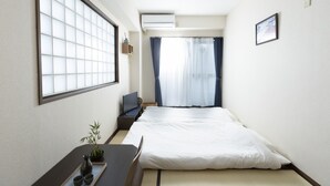 Een hotelkamer met een bed, een bureau met een stoel, een kleine tafel met een plant en kopjes, een televisie en een raam met gordijnen.