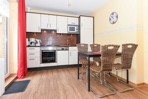 A compact kitchen with white cabinets, a microwave, and a coffee maker. There is a dining table with wicker chairs and a clock on the wall.