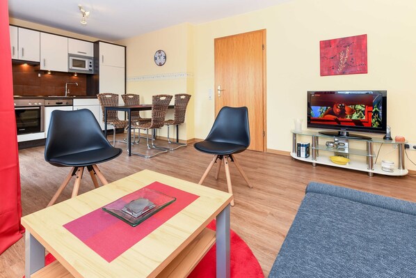 A modern living room with a TV, two black chairs, a wooden table, and a kitchen area with cabinets and appliances.