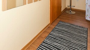 Une pièce comprenant une armoire en bois, un miroir, une petite fenêtre donnant sur l’extérieur, un tapis rayé noir et blanc et un petit tabouret.