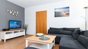 Un salon moderne avec un canapé gris, une table basse en bois, un téléviseur posé sur un meuble blanc et une porte.
