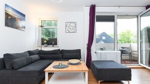 Un salon moderne avec un canapé d’angle, une table basse en bois et un pouf gris. Une fenêtre offre une vue sur la verdure et un bâtiment à l’extérieur.