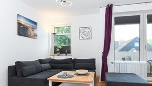 Un salon moderne avec un canapé d’angle gris foncé, une table basse en bois et un fauteuil gris. Une fenêtre offre une vue sur la verdure, et il y a un radiateur.