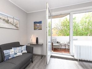 Ein modernes Wohnzimmer mit einem grauen Sofa, einem kleinen Tisch mit einer Lampe und einer Glastür, die auf eine Terrassenanlage mit Korbmöbeln führt.