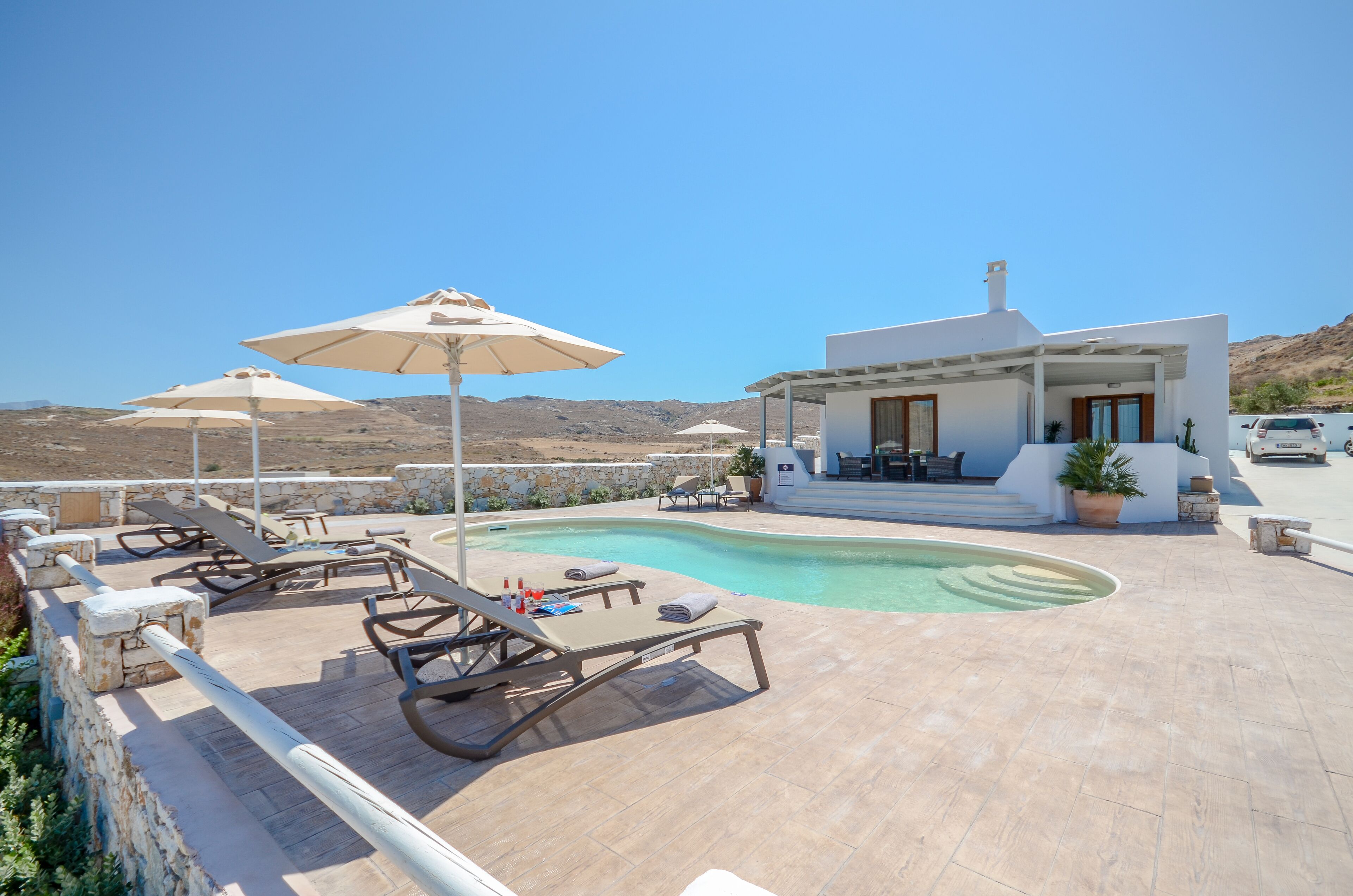 Un área de piscina con sillones de descanso y sombrillas, un edificio moderno con una zona de estar cubierta, y montañas al fondo.