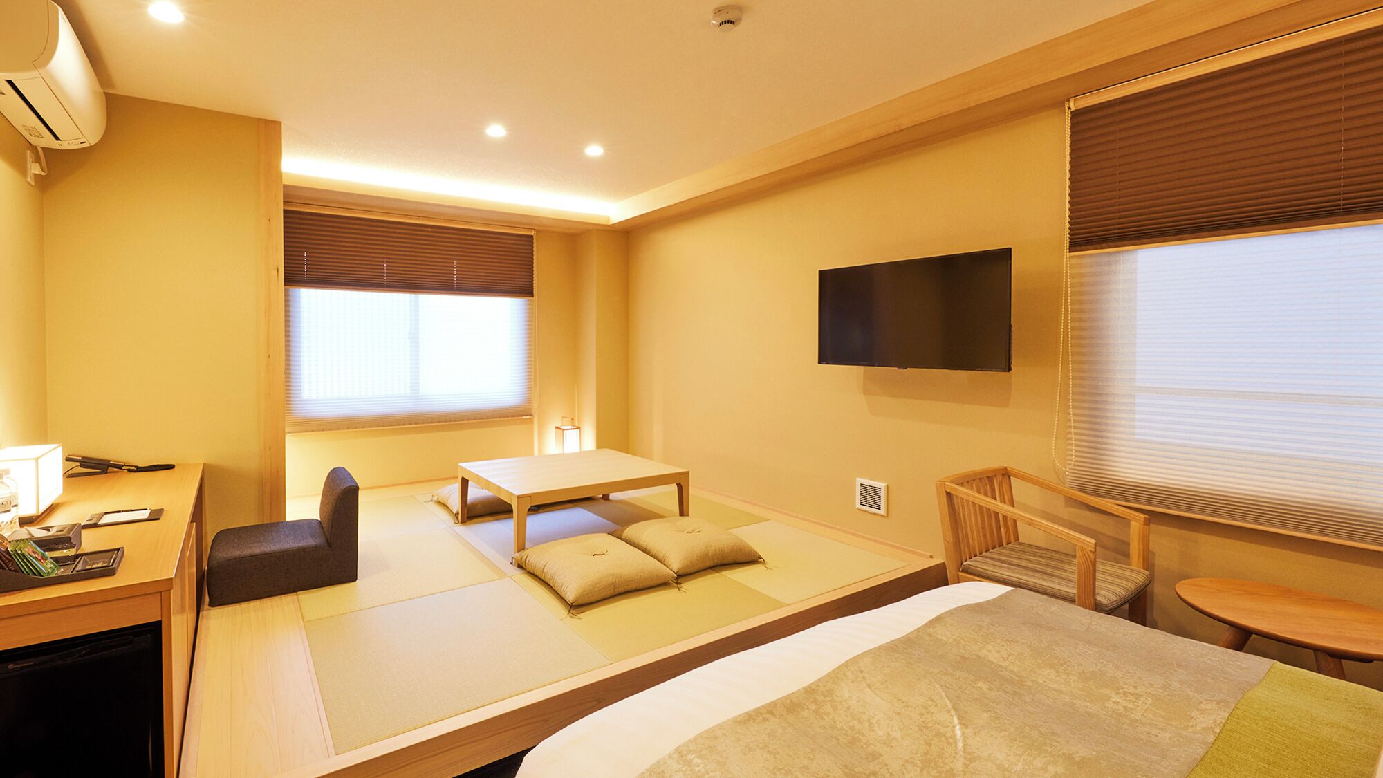 A hotel room with tatami flooring, a desk with a chair, a bed, and a TV mounted on the wall.