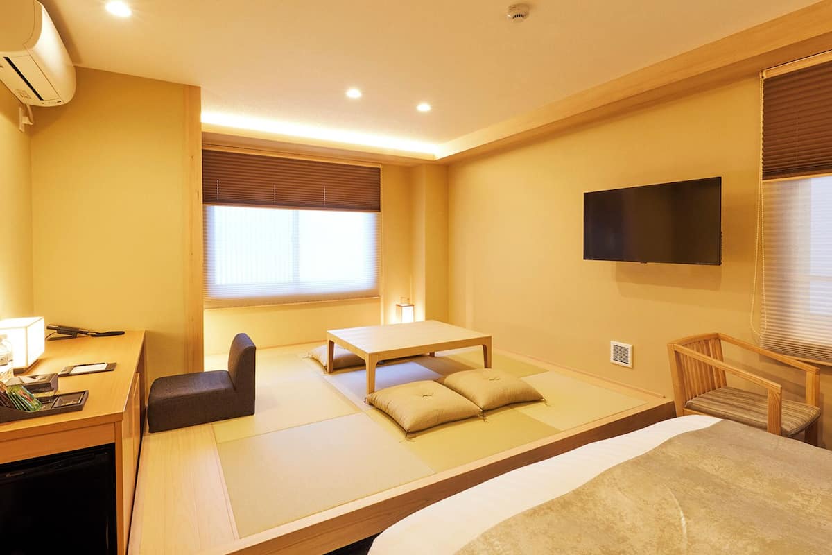 A hotel room with tatami flooring, a desk with a chair, a bed, and a TV mounted on the wall.