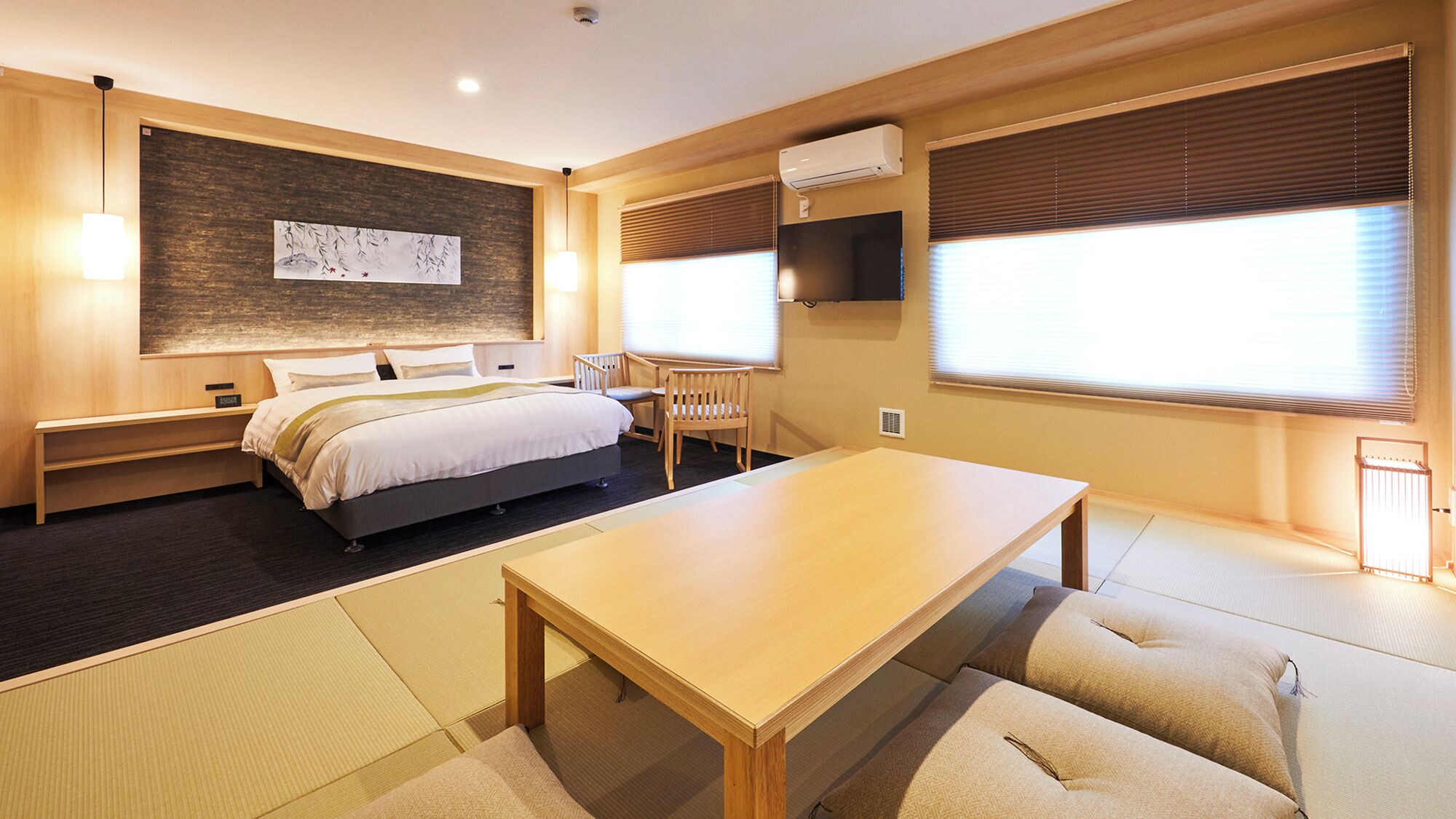 A hotel room with a bed, bedside table, chair, table, tatami mats, artwork, air conditioning unit, and windows with blinds.