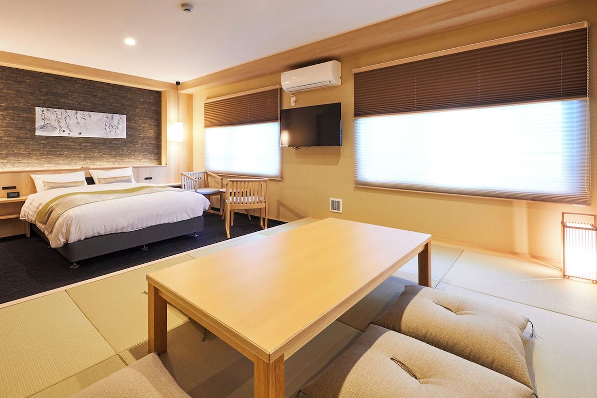 A hotel room with a bed, bedside table, chair, table, tatami mats, artwork, air conditioning unit, and windows with blinds.