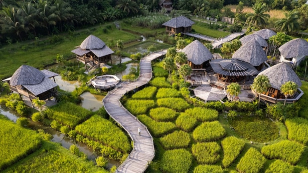 Vista aérea de un resort con cabañas de techo de paja conectadas por pasarelas de madera, rodeado de exuberante vegetación y cuerpos de agua.