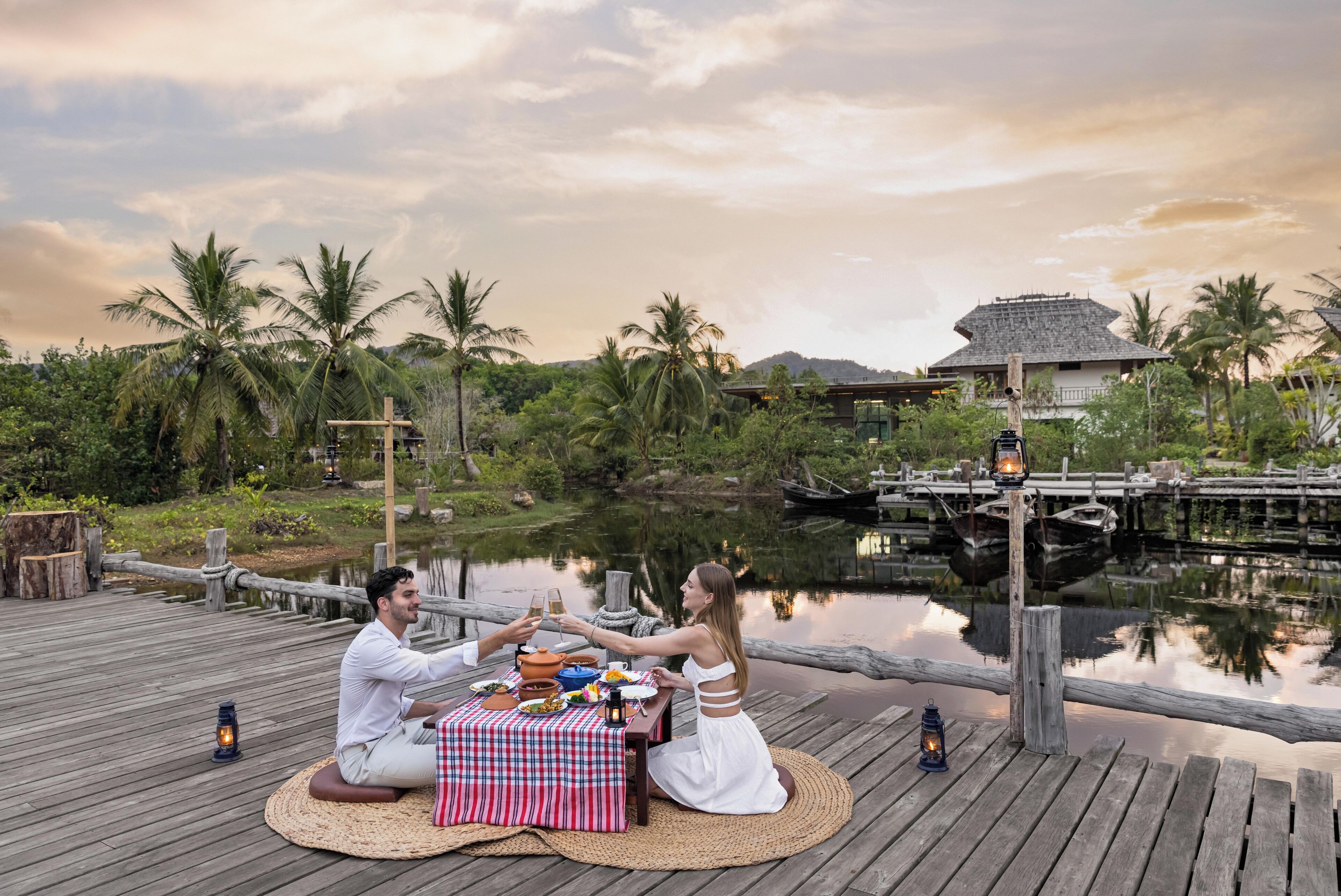 A couple toasting with drinks on a wooden deck by a calm lake, surrounded by lush greenery and a traditional hut in the background.