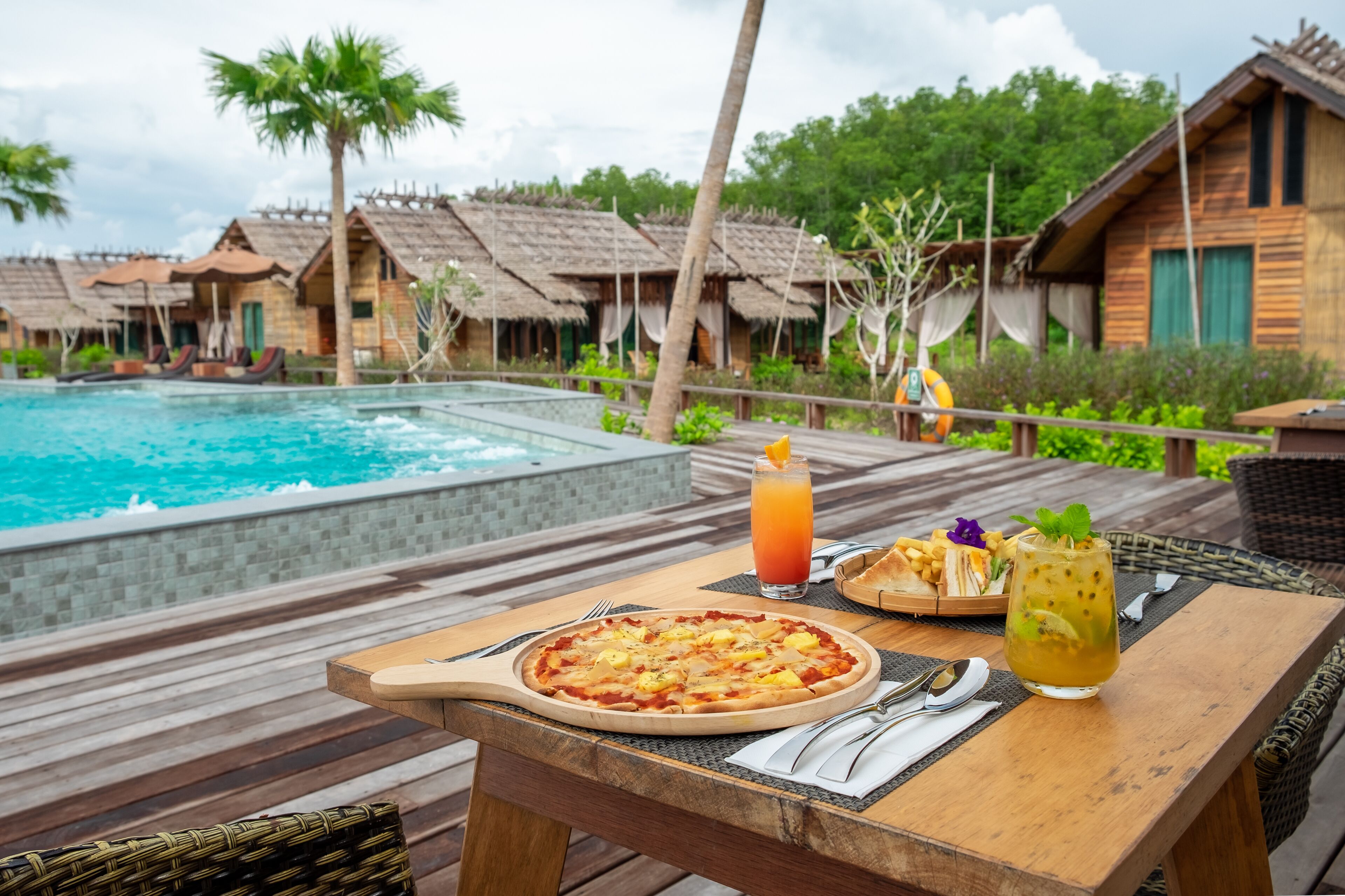 Un tavolo di legno con una pizza, un bicchiere di tè freddo e una bevanda con una fetta d'arancia, su una terrazza affacciata su una piscina e edifici con tetti di paglia.